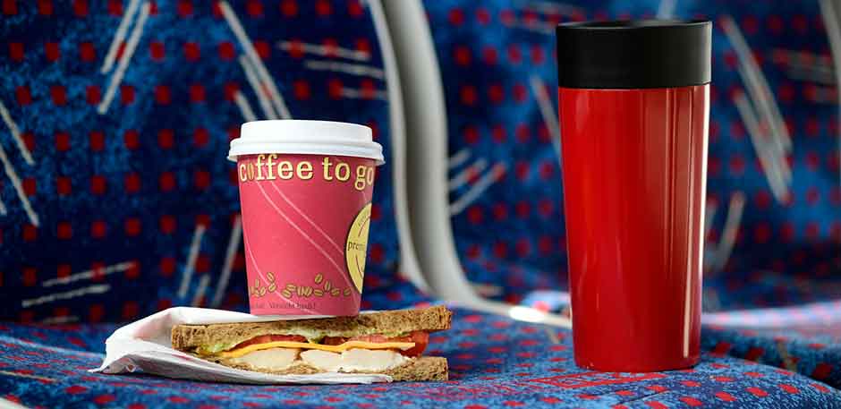 Sitze in Straßenbahn, auf denen ein Brot liegt und ein Coffee-to-go-Becher aus Pappe sowie ein Thermosbecher steht.