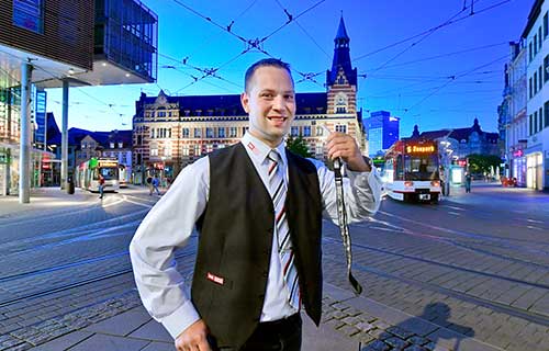 Straßenbahnfahrer mit Trillerpfeife in der Hand steht lächelnd auf dem Anger. Im Hintergrund ist fährt eine Straßenbahn vorbei.