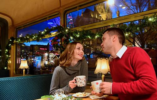 Mann und Frau sitzen im weihnachtlich geschm&uuml;ckten historischen Fahrzeug. Vor ihnen stehen Tassen und Geb&auml;ck. Im Hintergrund der weihnachtliche Domplatz.