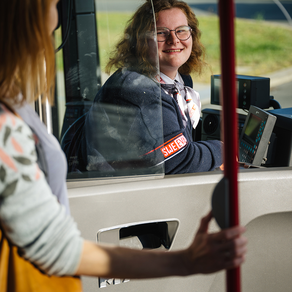 Busfahrerin sitzt in Fahrerkabine. Links steht eine junge Frau und drückt den Haltewunschtaster.