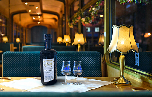 Stimmungsvoll gedeckter Tisch in einer nostalgischen Stra&szlig;enbahn mit einer Flasche Lik&ouml;r, zwei gef&uuml;llten Gl&auml;sern und einer warm leuchtenden Tischlampe im Hintergrund.