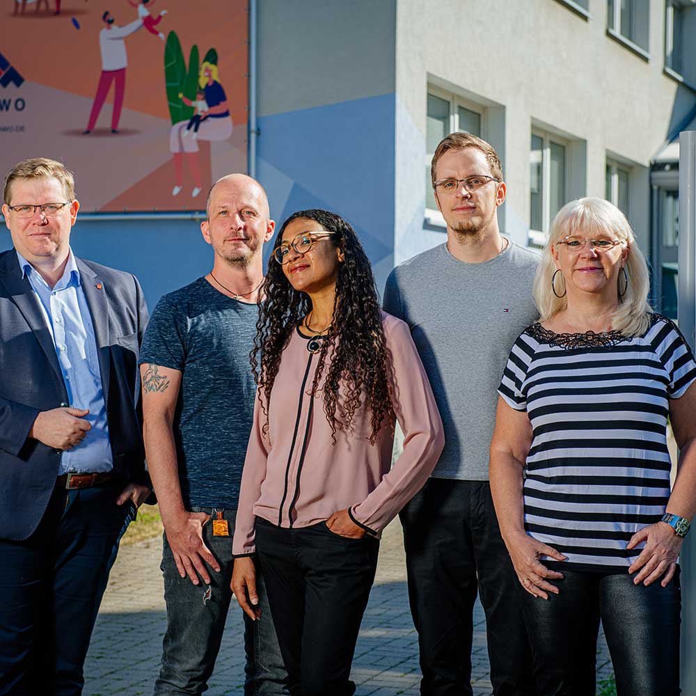 Drei M&auml;nner und zwei Frauen stehen vor einem blau bemalten Wohnblock.
