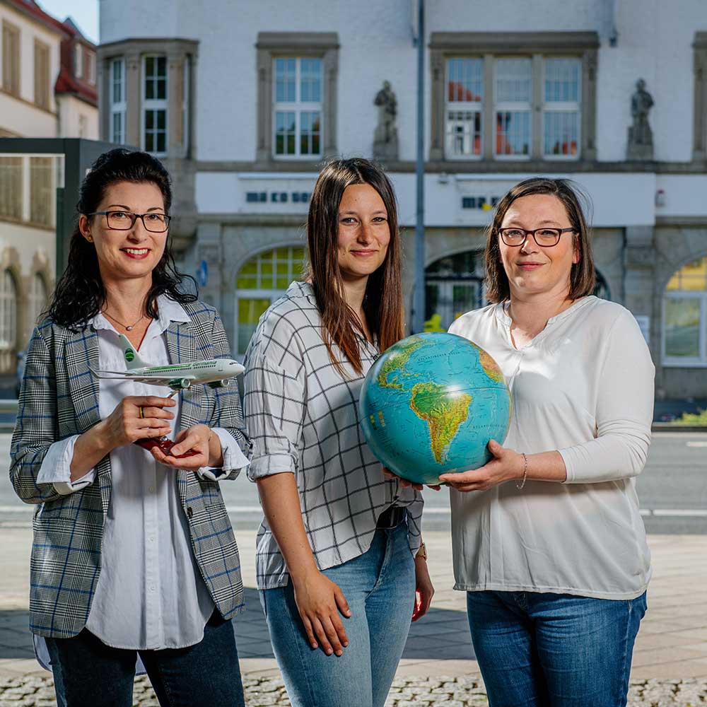 Drei Frauen stehen vor einem Gebäude. Eine hält einen Globus, eine andere ein Modellflugzeug.
