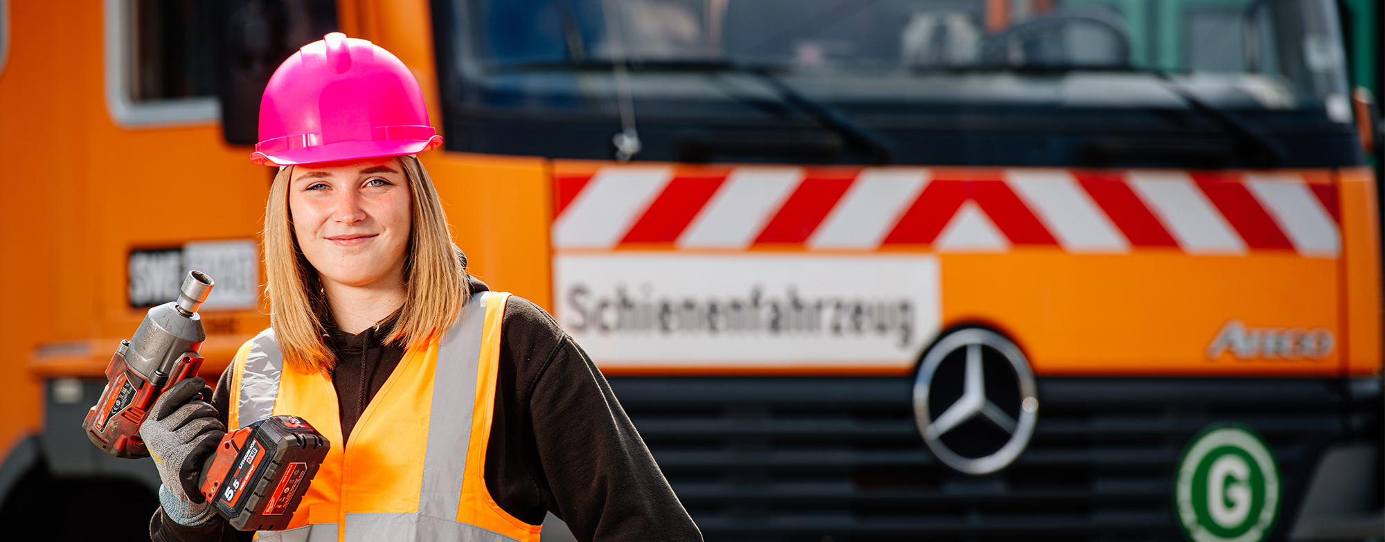 Mitarbeiterin mit Schlagbohrer in der Hand, Warnweste und pinkem Bauhelm.