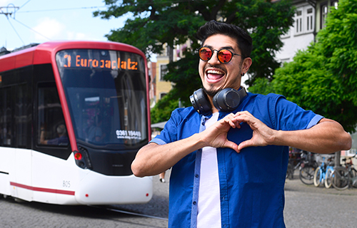 Junger Mann mit Sonnenbrille steht vor einer Stra&szlig;enbahn und formt mit den H&auml;nden ein Herz.