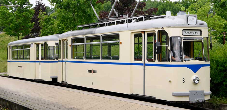 Historische Straßenbahn Nummer 3 in beiger Lackierung mit blauem Streifen in der Wendeschleife. Im Hintergrund grüne Bäume.