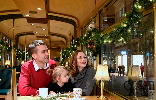 Mann, Frau und Tochter sitzen im weihnachtlich geschm&uuml;ckten historischen Fahrzeug. Vor ihnen stehen Tassen und Geb&auml;ck.