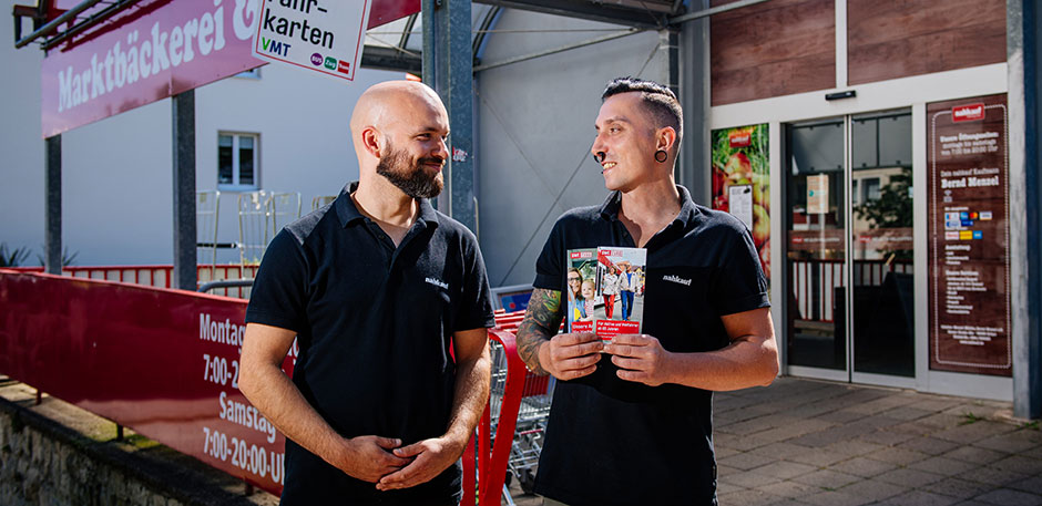 Zwei M&auml;nner stehen vor dem Eingang eines Supermarktes. Ein Mann h&auml;lt Flyer in den H&auml;nden.