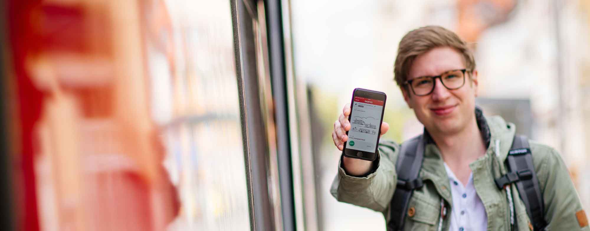 Fahrgast zeigt Handy mit FAIRTIQ-App auf dem Bildschirm. Rechts ist eine Stra&szlig;enbahn zu sehen. 