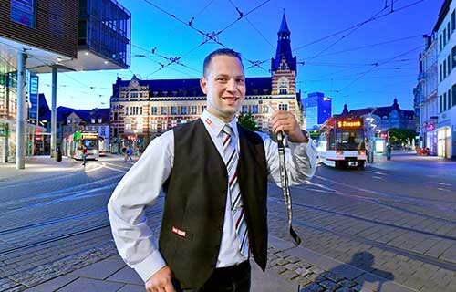 Stra&szlig;enbahnfahrer mit Trillerpfeife in der Hand steht l&auml;chelnd auf dem Anger. Im Hintergrund ist f&auml;hrt eine Stra&szlig;enbahn vorbei.