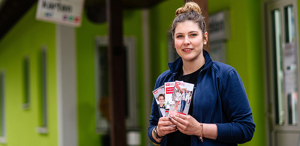 Eine Frau steht vor dem gr&uuml;n gestrichenen Empfangsgeb&auml;ude eines Campingplatzes. In der Hand h&auml;lt sie Fyer.