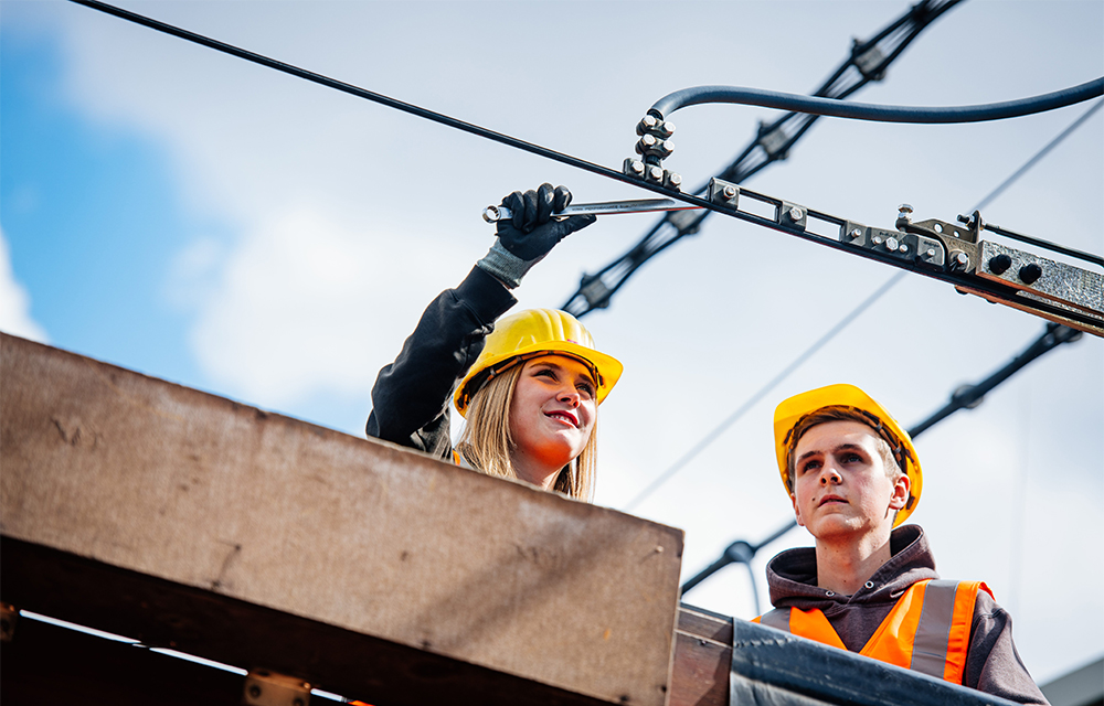 Zwei Azubi stehen auf einer Hebeb&uuml;hne unter einer Stromleitung.