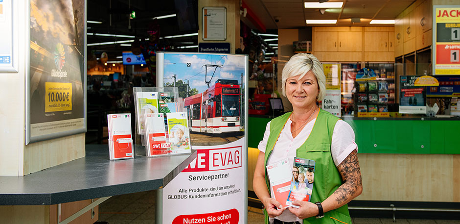 Frau steht mit Flyern in der Hand vor dem Infoschalter eines Supermarktes.